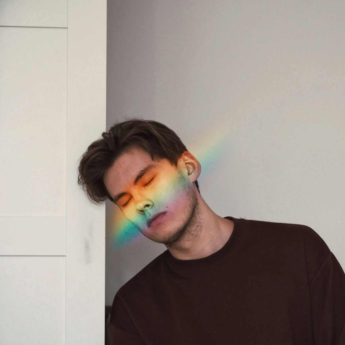 A person with eyes closed resting against a wall, with a rainbow light across their face, reflecting the personal impact of PTSD in LGBTQ lives.